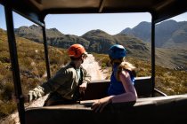 Vista lateral de la pareja caucásica disfrutando del tiempo en la naturaleza juntos, en equipo de forro de cremallera sentado en un coche, mirando hacia atrás en un día soleado en las montañas - foto de stock