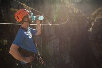 Side view of Caucasian man enjoying time in nature, wearing zip lining equipment, putting helmet on, drinking water, on a sunny day in mountains — Stock Photo
