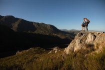Vue latérale du couple caucasien profitant du temps dans la nature ensemble, portant un équipement de tyrolienne, randonnée, debout sur un rocher, câlins, par une journée ensoleillée dans les montagnes — Photo de stock