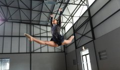 Низкий угол обзора подростковой смешанной расы гимнастки, выступающей в тренажерном зале, прыгающей и делающей разрыв, прыгающей и делающей разрыв, одетой в черно-фиолетовый трико — стоковое фото
