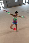 Vista frontal de ángulo alto de la gimnasta femenina adolescente de raza mixta que actúa en el gimnasio, haciendo ejercicio con una bola roja, de pie con la pelota entre sus pies, los brazos estirados, con maillot multicolor - foto de stock