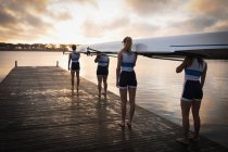 Передній вигляд веслувальної групи з чотирьох кавказьких жінок, які несуть човен на плечах, стоячи на пристані в річці на світанку. — стокове фото