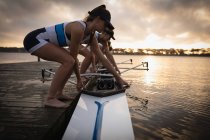 Vue latérale rapprochée d'une équipe d'aviron de quatre femmes caucasiennes debout sur une jetée et plaçant un bateau dans la rivière en préparation pour l'entraînement à l'aviron — Photo de stock
