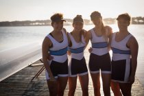 Retrato de uma equipe de remo de quatro mulheres caucasianas treinando no rio, de pé em um molhe com os braços ao redor um do outro, sorrindo para a câmera, um barco ao lado deles em um suporte — Fotografia de Stock