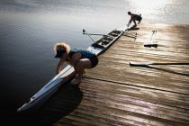 Vista lateral de dos mujeres remadoras caucásicas desde un equipo de remo entrenando en el río, de pie en un embarcadero y bajando un bote al agua - foto de stock