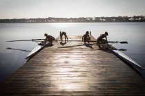 Seitenansicht eines Ruderteams von vier kaukasischen Frauen, die auf dem Fluss trainieren, auf einem Steg bei Sonnenaufgang, der zwei Boote vorbereitet, bevor sie rudern — Stockfoto