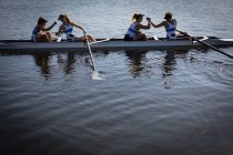 Vista lateral de un equipo de remo de cuatro mujeres caucásicas entrenando en el río, sentadas en una concha de carreras al sol dándose la mano y sonriendo después de remar - foto de stock