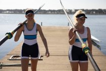 Front view of two Caucasian female rowers from a rowing team walking on a jetty in the sun carrying oars on their shoulders and smiling — Stock Photo