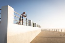 Low angle side view of a mature senior Caucasian man working out on a promenade on a sunny day with blue sky, taking a break, standing, holding water bottle, leaning on a balustrade — Stock Photo