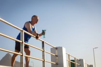 Vue latérale à angle bas d'un homme caucasien âgé mature travaillant sur une promenade par une journée ensoleillée avec un ciel bleu, prenant une pause, debout, tenant une bouteille d'eau, s'appuyant sur une balustrade — Photo de stock