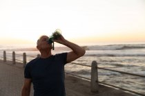 Vorderansicht eines älteren kaukasischen Mannes, der an einem sonnigen Tag auf einer Promenade trainiert, eine Pause macht, steht und aus einer Wasserflasche trinkt — Stockfoto