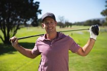 Портрет кавказького чоловіка на полі для гольфу в сонячний день з блакитним небом, тримаючи гольф клуб через плечі, посміхаючись до камери — стокове фото