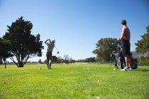 Вид сзади на двух белых мужчин на поле для гольфа в солнечный день с голубым небом, один бьет по мячу, а другой стоит и смотрит — стоковое фото