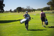 Задній вигляд двох кавказьких чоловіків на полі для гольфу в сонячний день з блакитним небом, ходити, несучи мішки для гольфу. — стокове фото