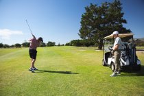 Вид сзади на двух белых мужчин на поле для гольфа в солнечный день с голубым небом, один бьет по мячу, а другой стоит и смотрит — стоковое фото