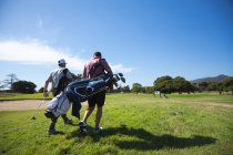 Задній вигляд двох кавказьких чоловіків на полі для гольфу в сонячний день з блакитним небом, ходити, несучи мішки для гольфу. — стокове фото