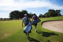 Rückansicht zweier kaukasischer Männer auf einem Golfplatz an einem sonnigen Tag mit blauem Himmel, beim Gehen, Golftaschen tragend — Stockfoto
