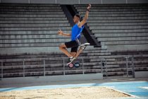 Vue latérale d'un athlète masculin de race mixte pratiquant dans un stade de sport, faisant un saut en longueur. Entraînement sportif d'athlétisme dans le stade. — Photo de stock