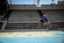 Vista lateral de um atleta misto praticando em um estádio de esportes, fazendo um salto em distância. — Fotografia de Stock
