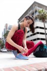 Front view of a fit Caucasian woman on her way to fitness training on a cloudy day, carrying a sports bag and a yoga mat, kneeling down in the street and tying her shoelaces — Stock Photo