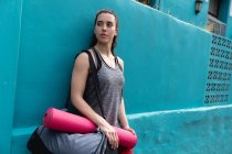 Vista frontale di una donna caucasica in forma sulla strada per l'allenamento fitness in una giornata nuvolosa, portando una borsa sportiva e un tappetino yoga, appoggiata a una parete blu — Foto stock