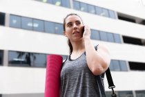 Front view of a fit Caucasian woman on her way to fitness training on a cloudy day, carrying a sports bag and a yoga mat, talking on a smartphone and walking in the street — Stock Photo