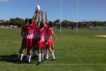 Vista trasera de un grupo de jugadores de rugby masculinos multiétnicos adolescentes que usan tira de equipo roja y blanca, de pie en un campo de juego y elevando la pelota de rugby en el aire - foto de stock
