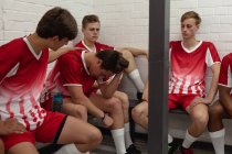 Seitenansicht einer Gruppe multiethnischer männlicher Rugby-Spieler im rot-weißen Mannschaftskleid, die nach einem Spiel in der Umkleidekabine sitzen und sich ausruhen, während ein Spieler seinem Teamkollegen auf die Schulter klopft. — Stockfoto
