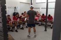 Vista trasera de un entrenador de rugby masculino caucásico de pie en el vestuario hablando con un equipo masculino multiétnico adolescente de jugadores de rugby que usan su tira de equipo, sentado y escuchando - foto de stock