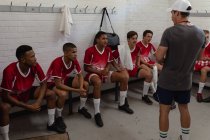 Rückansicht eines kaukasischen männlichen Rugby-Trainers, der in der Umkleidekabine steht und mit einer multiethnischen, männlichen Rugby-Mannschaft spricht, die ihre Mannschaftskleidung trägt, sitzt und zuhört — Stockfoto