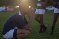 Side view of a teenage mixed-race male rugby player wearing blue and white team strip, squatting on a playing field, resting after a rugby match, with other players in behind, backlit — Stock Photo