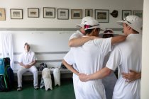 Vista lateral de um grupo de jogadores de críquete masculinos multi-étnicos adolescentes vestindo brancos, abraçando em um vestiário, com outro jogador descansando em um banco. — Fotografia de Stock