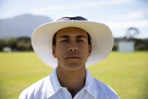 Portrait of a confident teenage Caucasian male cricket player wearing cricket whites, and a wide brimmed hat and sunglasses, standing on a cricket pitch on a sunny day looking to camera — Stock Photo