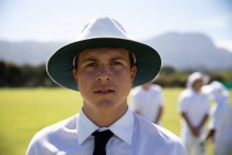Portrait d'un arbitre caucasien confiant portant une chemise blanche, une cravate noire et un chapeau à large bord, debout sur un terrain de cricket par une journée ensoleillée regardant vers la caméra, avec des joueurs de cricket debout derrière. — Photo de stock