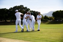 Вид сбоку на юношескую многонациональную команду по крикету в белых одеждах, стоящую на поле, обнимающуюся, улыбающуюся и разговаривающую в солнечный день. — стоковое фото