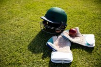 Feche a visão de alto ângulo de uma bola de críquete vermelha, um capacete de críquete verde e luvas de críquete deitado em um campo de críquete em um dia ensolarado — Fotografia de Stock