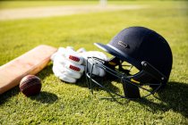 Vista de cerca de una pelota de cricket roja, un casco de cricket verde, un bate de cricket y guantes de cricket acostados en un campo de cricket en un día soleado - foto de stock