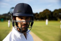 Porträt eines selbstbewussten, gemischten Cricket-Spielers mit Cricket-Weiß und Helm, der an einem sonnigen Tag auf einem Cricketplatz steht und in die Kamera blickt, während andere Spieler im Hintergrund stehen. — Stockfoto