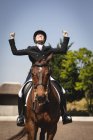 Vista frontale di una cavallerizza caucasica vestita elegantemente seduta su un cavallo di castagno in un recinto, che alza le mani e sorride in festa durante una gara di dressage in una giornata di sole. — Foto stock