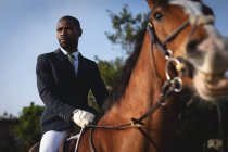 Vista frontale da vicino di un uomo afroamericano elegantemente vestito seduto su un cavallo di castagno durante una giornata di sole. — Foto stock