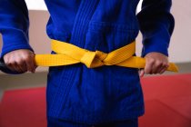 Front view mid section of judoka standing on gym mats, tying up the yellow belt of blue judogi. — Stock Photo
