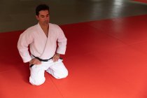 Vista frontal de um treinador de judô masculino de raça mista focado vestindo judogi branco, ajoelhado em esteiras no ginásio antes do treinamento de judô. — Fotografia de Stock