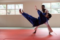 Vista lateral de un entrenador de judo masculino de raza mixta y judoka masculino de raza mixta adolescente, usando judogi azul y blanco, practicando judo durante un entrenamiento en un gimnasio. - foto de stock