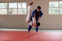 Vue latérale d'un entraîneur de judo masculin de race mixte et d'un judoka masculin de race mixte adolescent, portant du judogi bleu et blanc, pratiquant le judo lors d'un entraînement dans une salle de gym. — Photo de stock