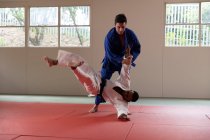 Vue latérale d'un entraîneur de judo masculin de race mixte et d'un judoka masculin de race mixte adolescent, portant du judogi bleu et blanc, pratiquant le judo lors d'un entraînement dans une salle de gym. — Photo de stock