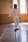 Desportista afro-americano vestindo roupa de esgrima protetora durante uma sessão de treinamento de esgrima, preparando-se para um duelo, segurando um epee. Treinamento de esgrimistas em um ginásio. — Fotografia de Stock