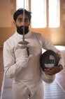 Retrato de esportista de raça mista vestindo roupa de esgrima protetora durante uma sessão de treinamento de esgrima, olhando para a câmera, segurando um epee. Treinamento de esgrimistas em um ginásio. — Fotografia de Stock