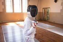 Caucasian sportswoman wearing protective fencing outfit during a fencing training session, preparing for a duel, holding an epee in front of face. Fencers training at a gym. — Stock Photo