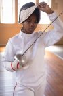 Desportista afro-americana vestindo roupa de esgrima protetora durante uma sessão de treinamento de esgrima, preparando-se para um duelo, segurando um epee e levantando sua máscara. Treinamento de esgrimistas em um ginásio. — Fotografia de Stock