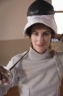 Portrait of Caucasian sportswoman wearing protective fencing outfit during a fencing training session, looking at camera with her mask raised and smiling, holding an epee. Fencers training at a gym. — Stock Photo